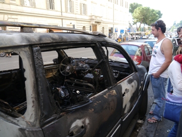 fiatal férfi áll egy felrobbantott, kiégett autó mellett