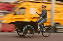 Nő biciklizik és közben sms-ezik, háttérben száguldó autó